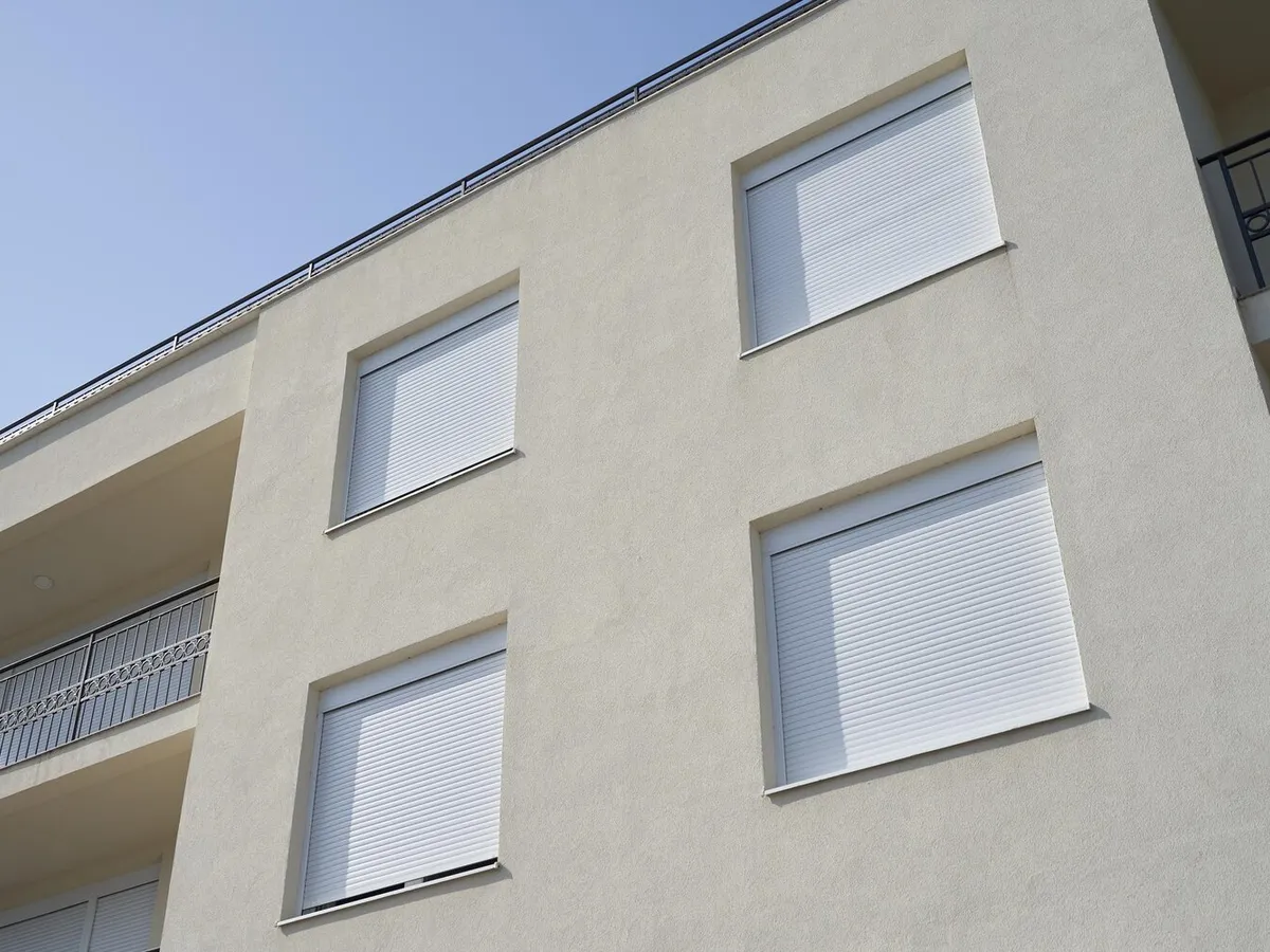 Façade d'immeuble à l'enduit clair dont les fenêtres sont équipées de volets roulants blancs à demi baissés, sous un ciel bleu
